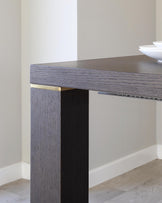 Modern minimalist dark wood dining table with a sleek design and subtle gold metal accents on the corners, set against a neutral backdrop with a white bowl on top.