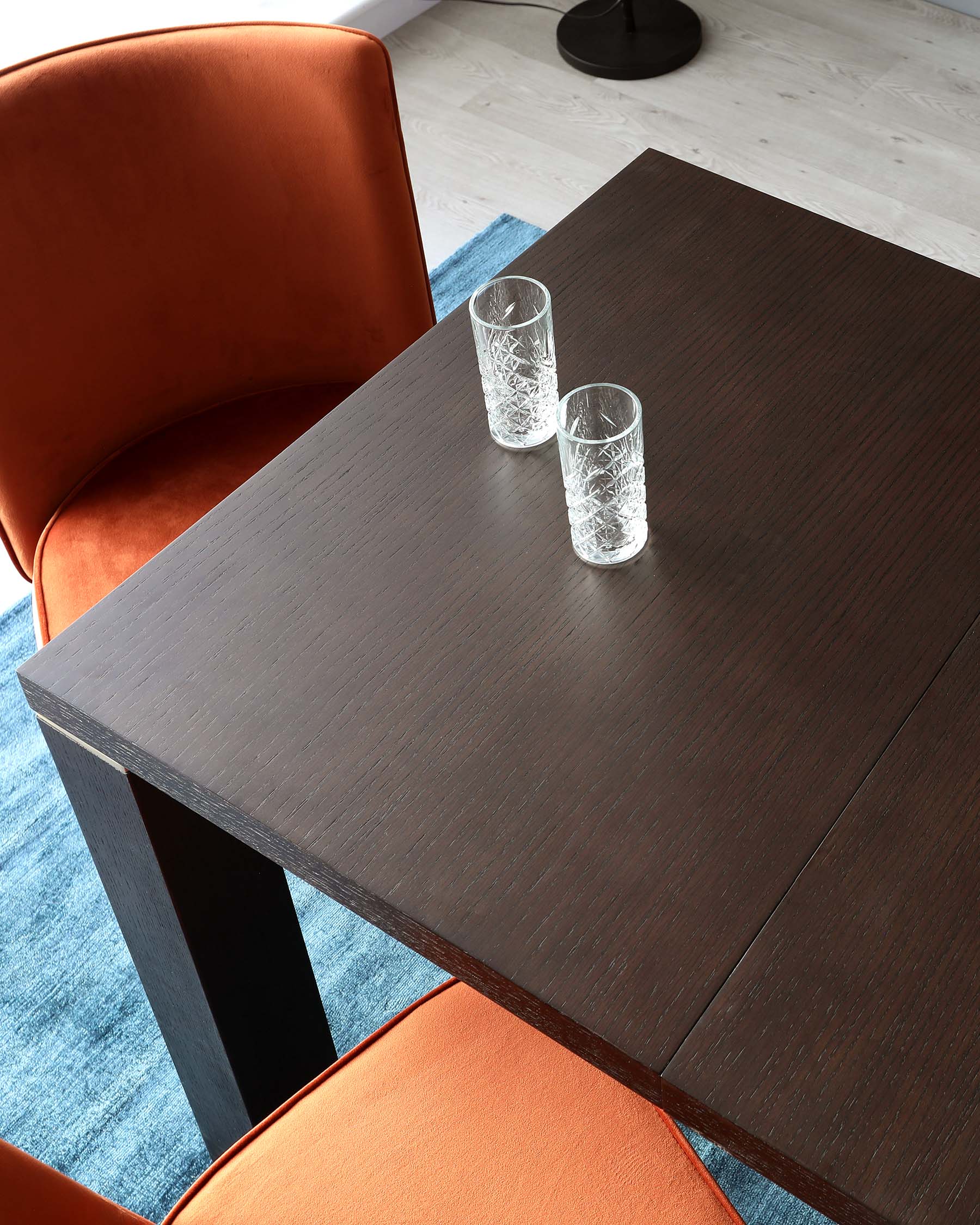 A modern rectangular dark wood dining table with a clean-lined design, accompanied by a curved, burnt orange upholstered dining chair with a smooth, supportive backrest. The table is adorned with two decorative cut-glass tumblers. The set is positioned on a light wood floor with a partial view of a blue area rug.