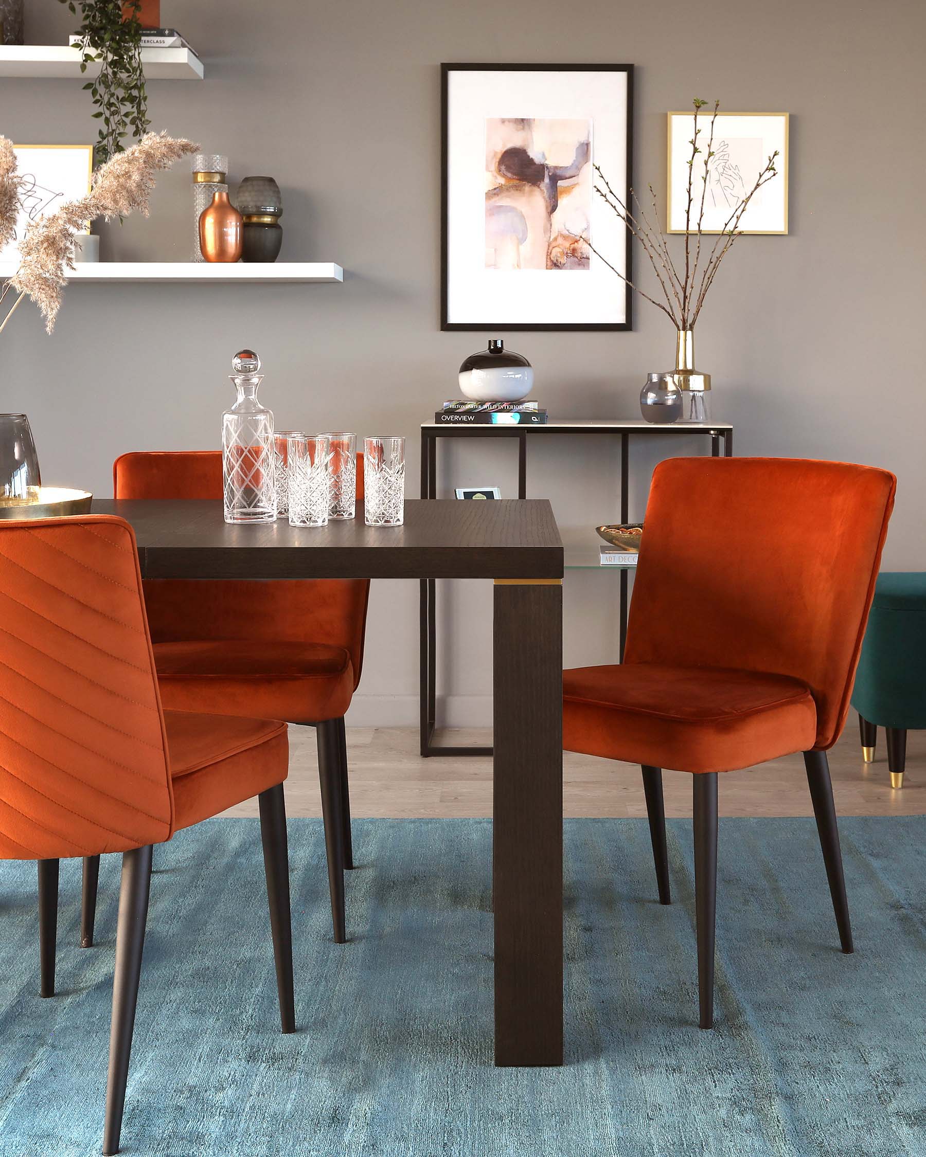 Modern dining space with a rectangular dark wood table featuring a minimalistic design and straight lines, complemented by two upholstered dining chairs in rich orange velvet with vertical stitching on the backrest and slender black metal legs. A sleek black console table with a lower shelf stands against the wall, displaying decorative items. The furniture is set on a soft blue rug, creating a cosy yet contemporary ambiance.