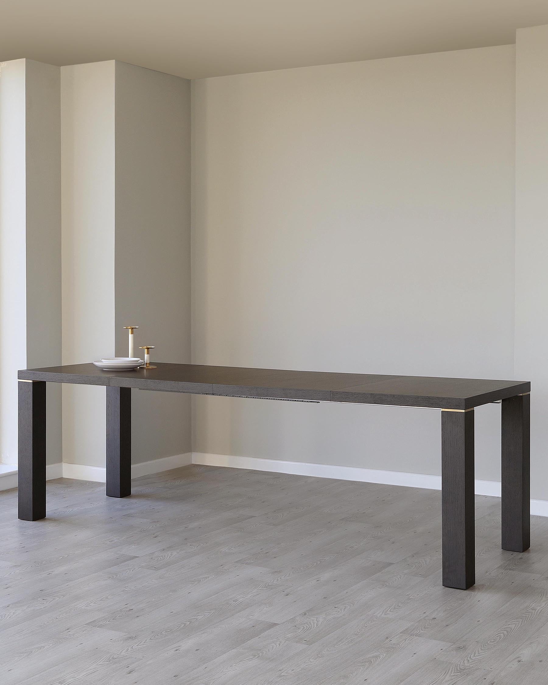 A modern minimalist rectangular dining table with a dark woodgrain finish and sturdy block legs, situated in an interior with light grey floors and neutral walls. On top of the table, there is a small white dish holding two gold-coloured candlesticks.