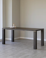 Modern minimalist dark wooden dining table with a rectangular top and four solid square legs. The table is accented with thin metallic details near the joints, and there are two small decorative items on its surface. The background reveals a room with light wooden flooring and pale walls, emphasizing the table's stark design.