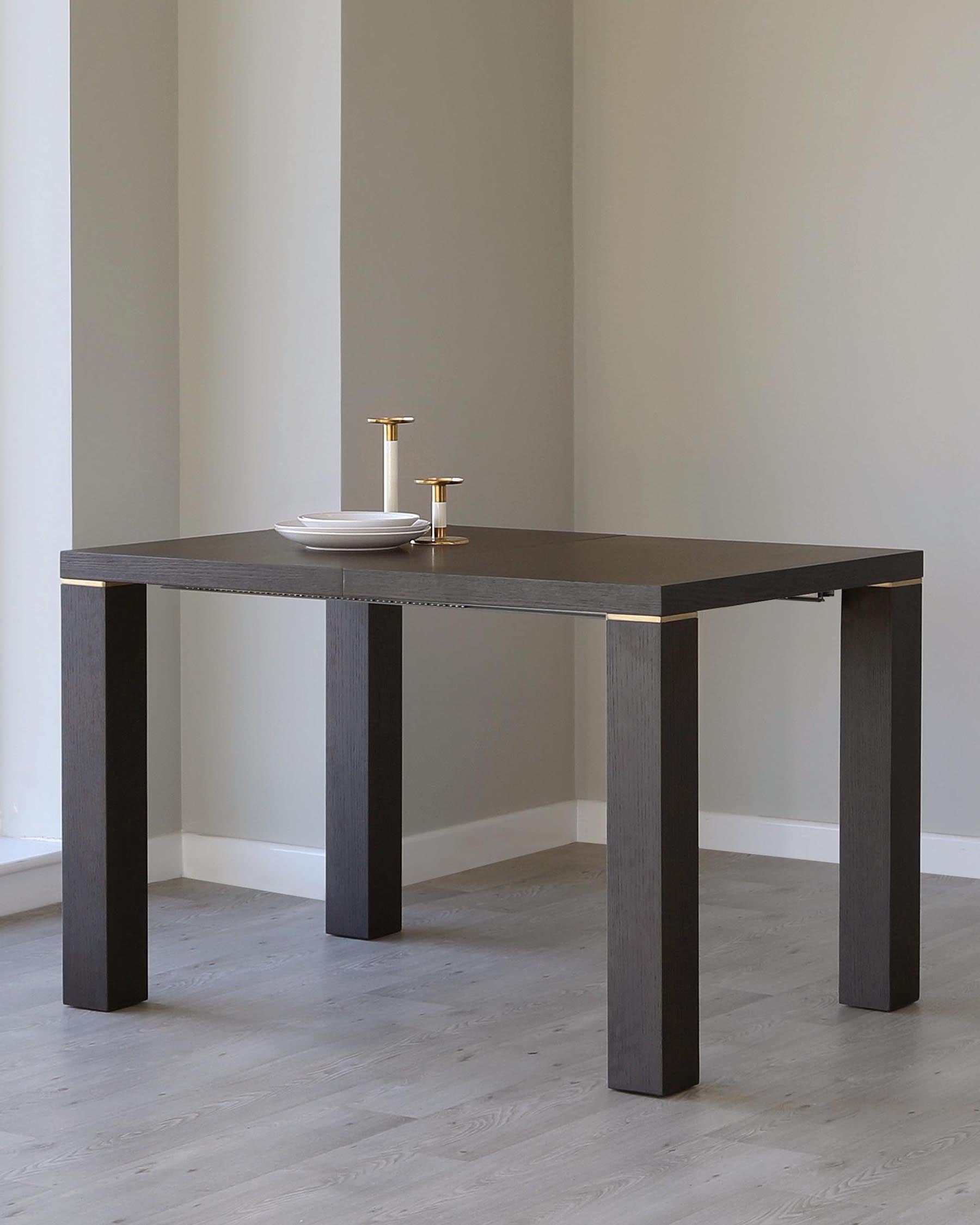 Modern minimalist dark wooden dining table with thick square legs, set against a neutral wall, adorned with a simple centrepiece of white plates and golden candlesticks.