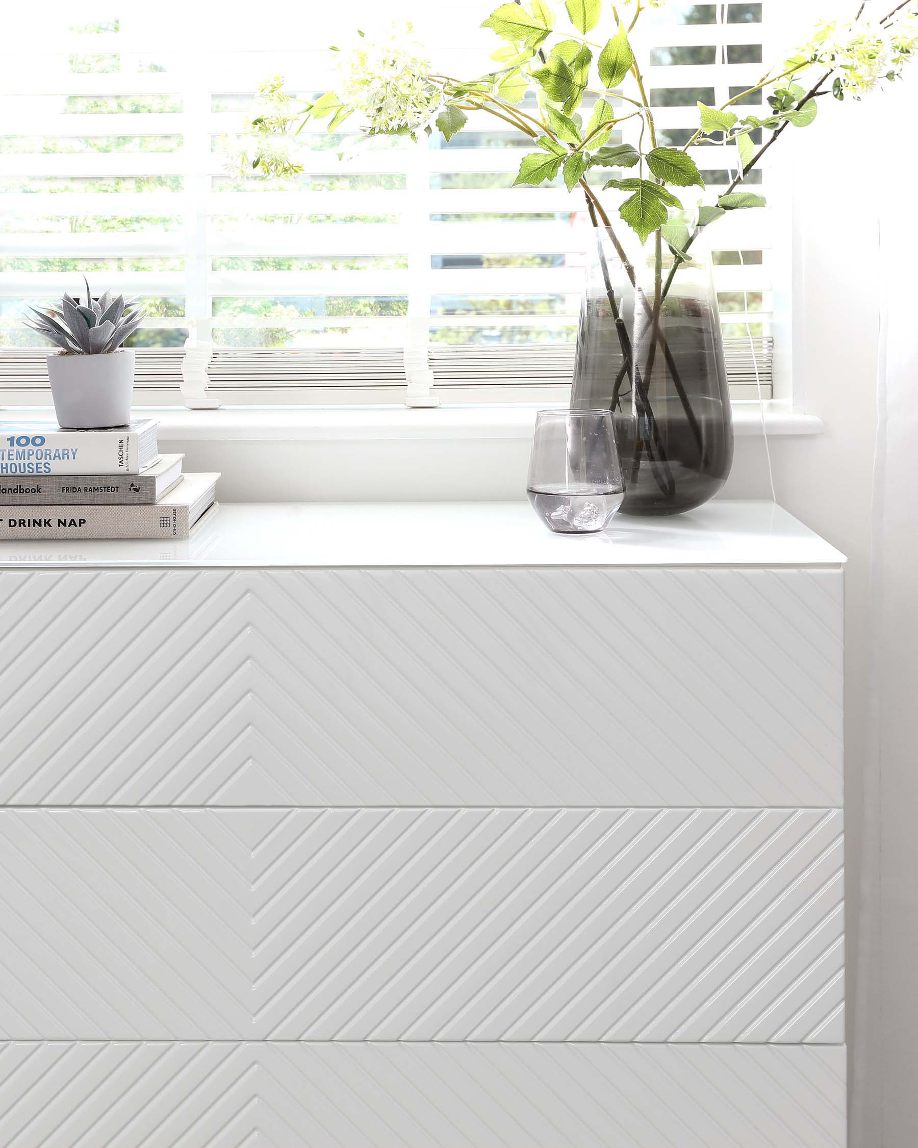 Modern white dresser with a distinctive chevron pattern on the drawers, set against a bright window with horizontal blinds.