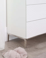 A modern white chest of drawers with geometric relief patterns on the drawer fronts and sleek metallic legs.