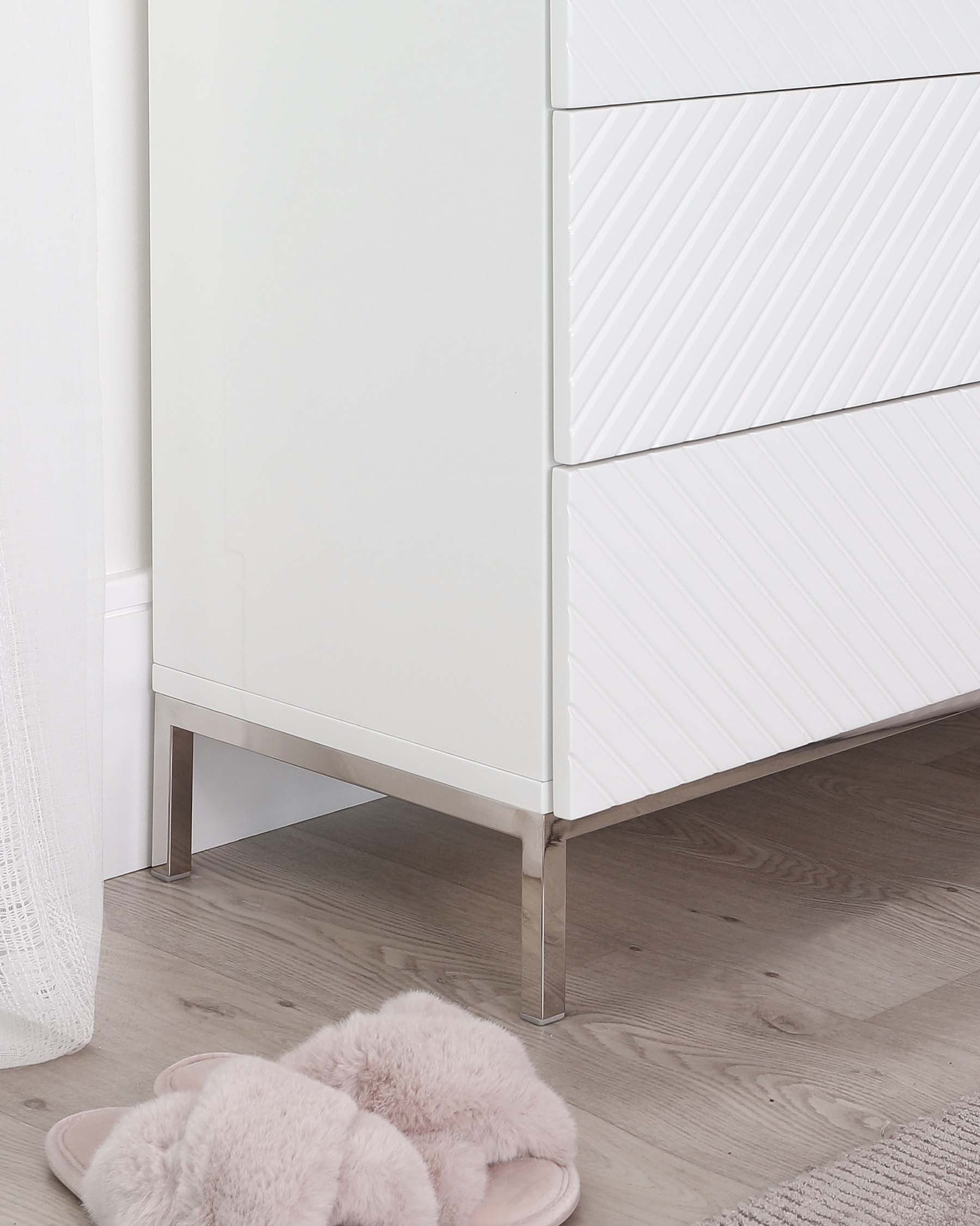 A modern white chest of drawers with geometric relief patterns on the drawer fronts and sleek metallic legs.