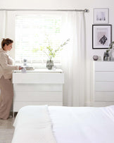 A modern bedroom featuring a white textured drawer dresser with a geometric pattern next to a bed with white bedding. A vase with fresh flowers sits atop the dresser, adding a touch of nature to the sleek and minimalist design.