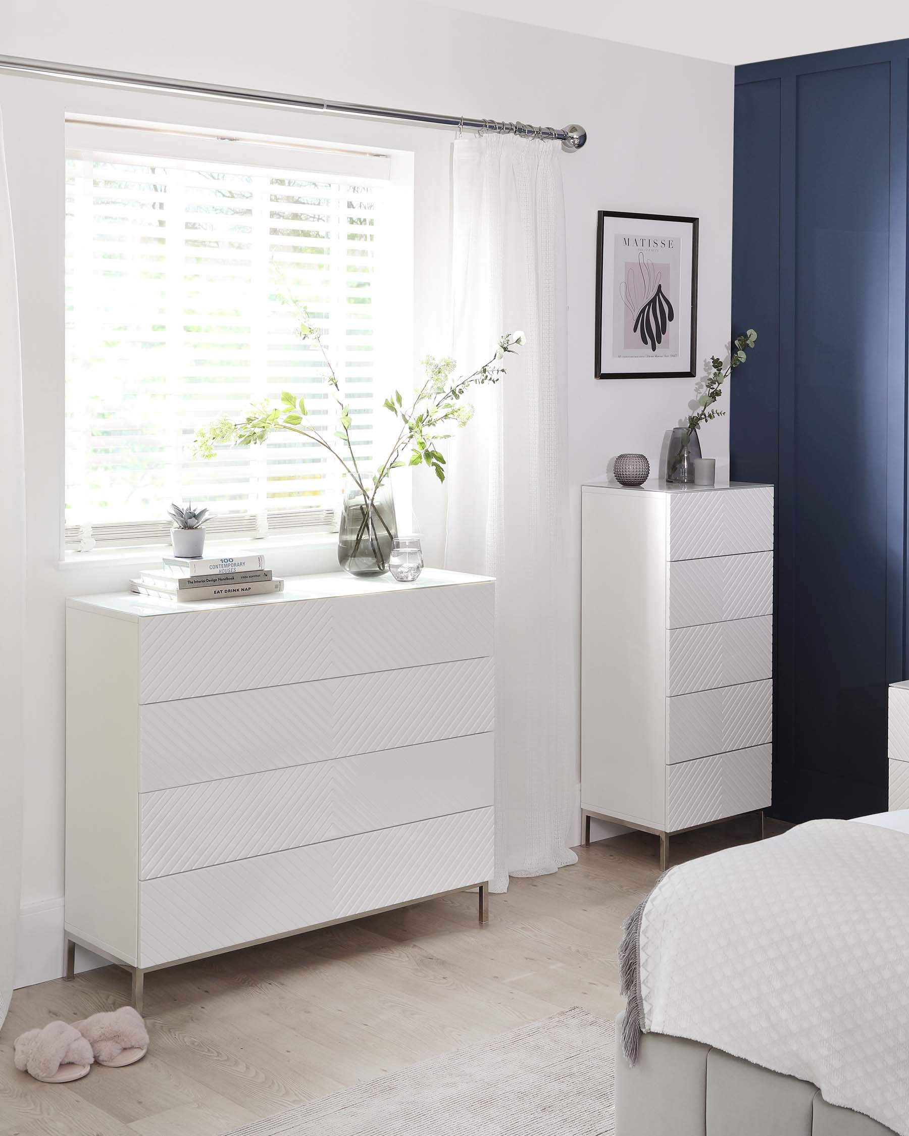 A contemporary white bedroom furniture set including a drawer dresser and a 5-drawer tall chest. Both pieces feature a modern geometric textured front, sleek metal legs, and minimalist handles.