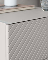 Modern light grey sideboard with a sleek finish and diagonally grooved front panels. The top surface holds a book and a glass of water, suggesting a functional yet stylish design suitable for contemporary living spaces.