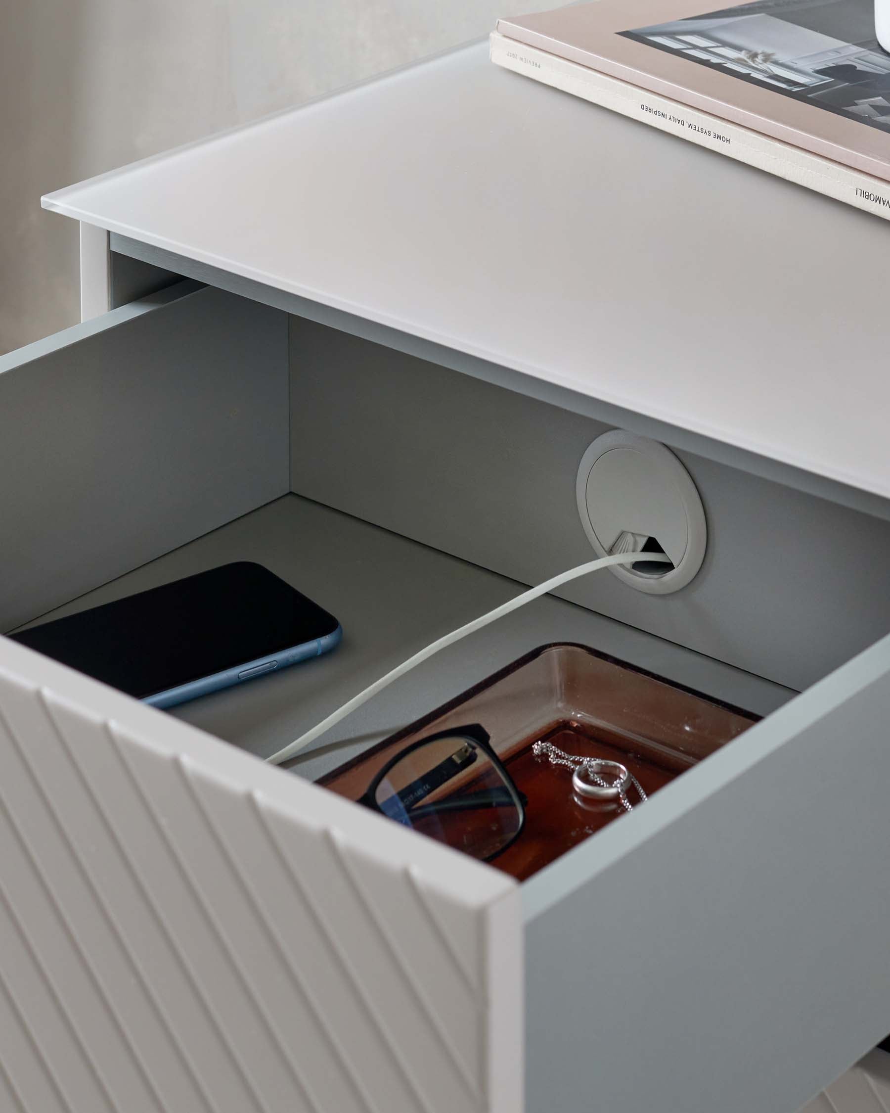 Modern minimalist nightstand with an open drawer showcasing a built-in organizer, featuring a grey finish with ribbed texturing on the drawer front, and an elegant white tabletop with a charging cable routed through a discreet hole.