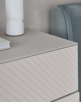 Modern minimalist nightstand with textured front in a light grey tone, paired with a sleek ceramic vase and a stack of hardcover books. In the background, a hint of a plush, velvet blue chair with a rounded backrest.