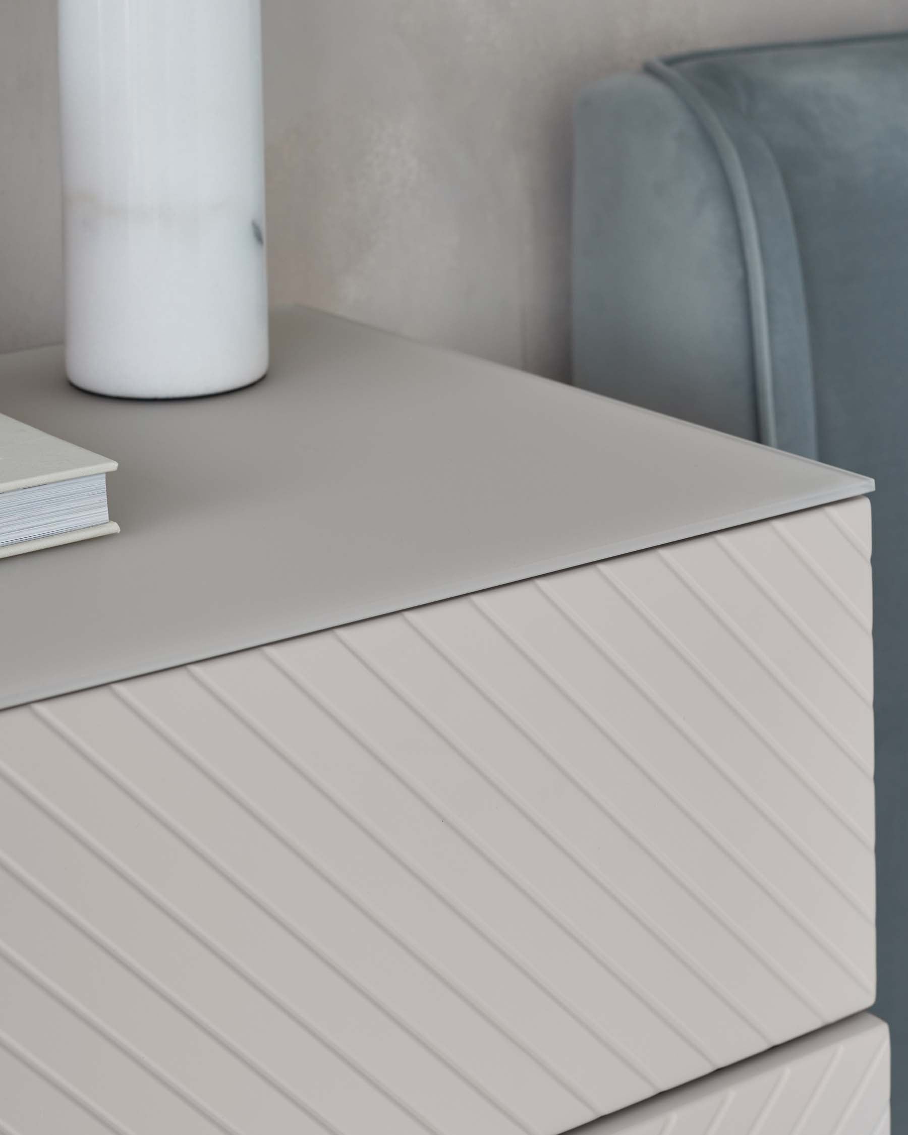 Modern minimalist nightstand with textured front in a light grey tone, paired with a sleek ceramic vase and a stack of hardcover books. In the background, a hint of a plush, velvet blue chair with a rounded backrest.