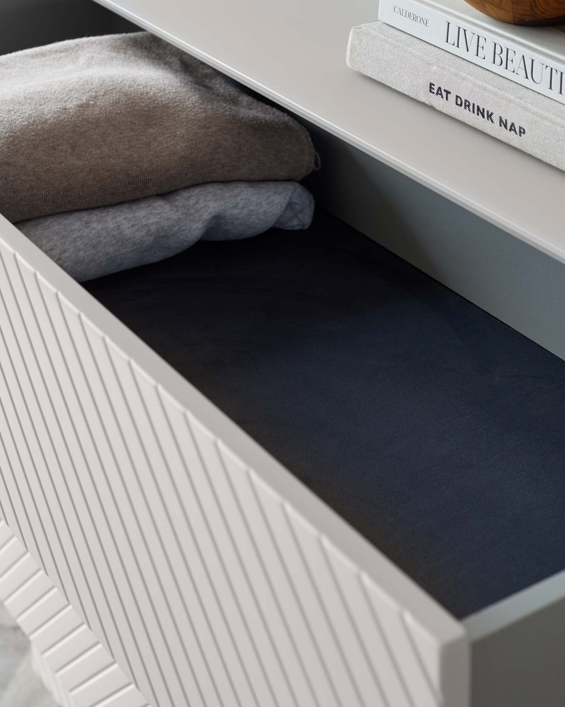 Close-up view of a modern grey storage bench with elegant vertical line detailing on the front panel, featuring an open compartment containing folded blankets. On top, there are two books stacked neatly to the side.