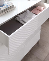 Modern white textured sideboard with an open drawer revealing interior storage space, positioned on light wooden legs, with books placed on top alongside a decorative glass item.