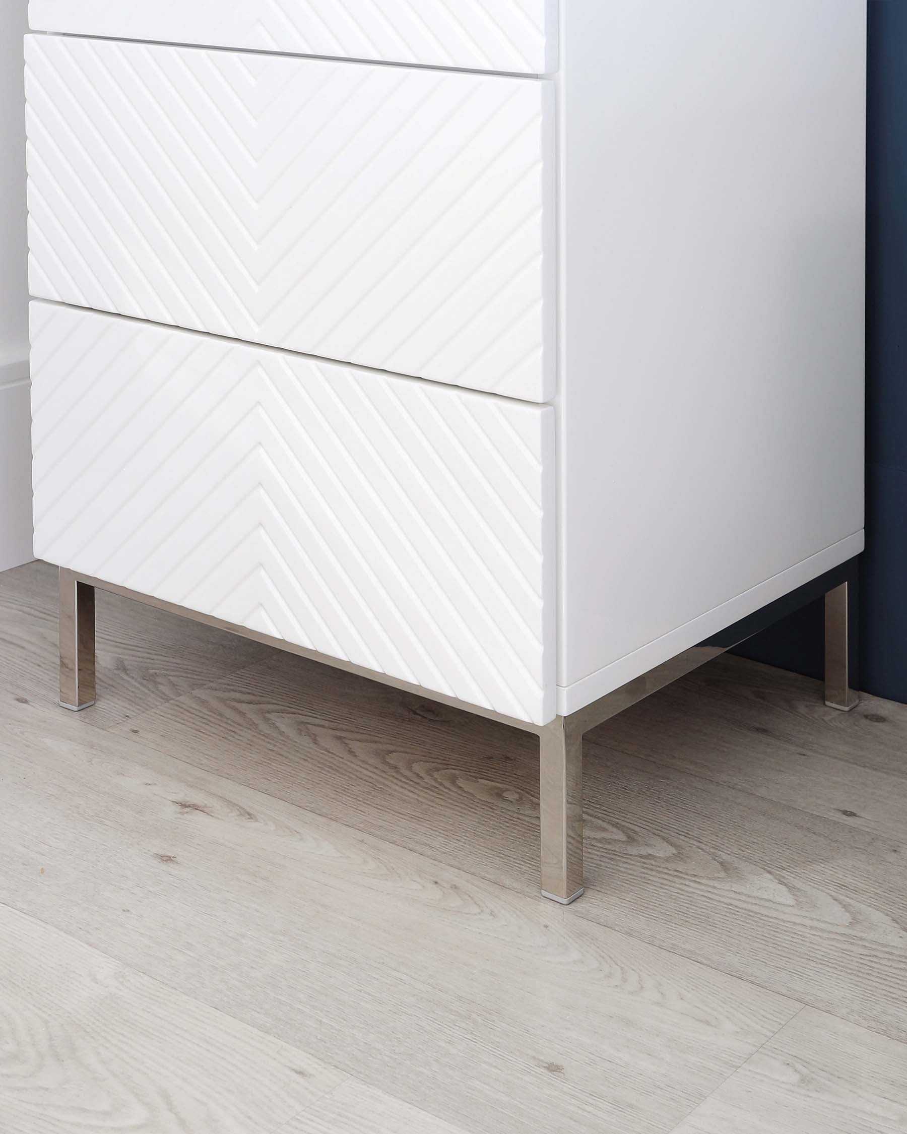 A modern white three-drawer dresser with a chevron pattern on the drawer fronts, poised on sleek metal legs, set against a light wood floor and wall with a dark blue accent.