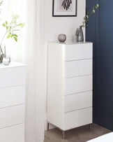 A contemporary white tallboy dresser with a geometric embossed pattern on the drawer fronts, featuring a narrow silhouette with five drawers and standing on sleek metallic legs.