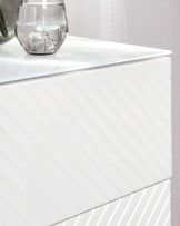 White modern chest of drawers with a high-gloss finish and a textured front displaying diagonal line patterns, accompanied by a clear glass vase and a glass with an ice cube on its surface.