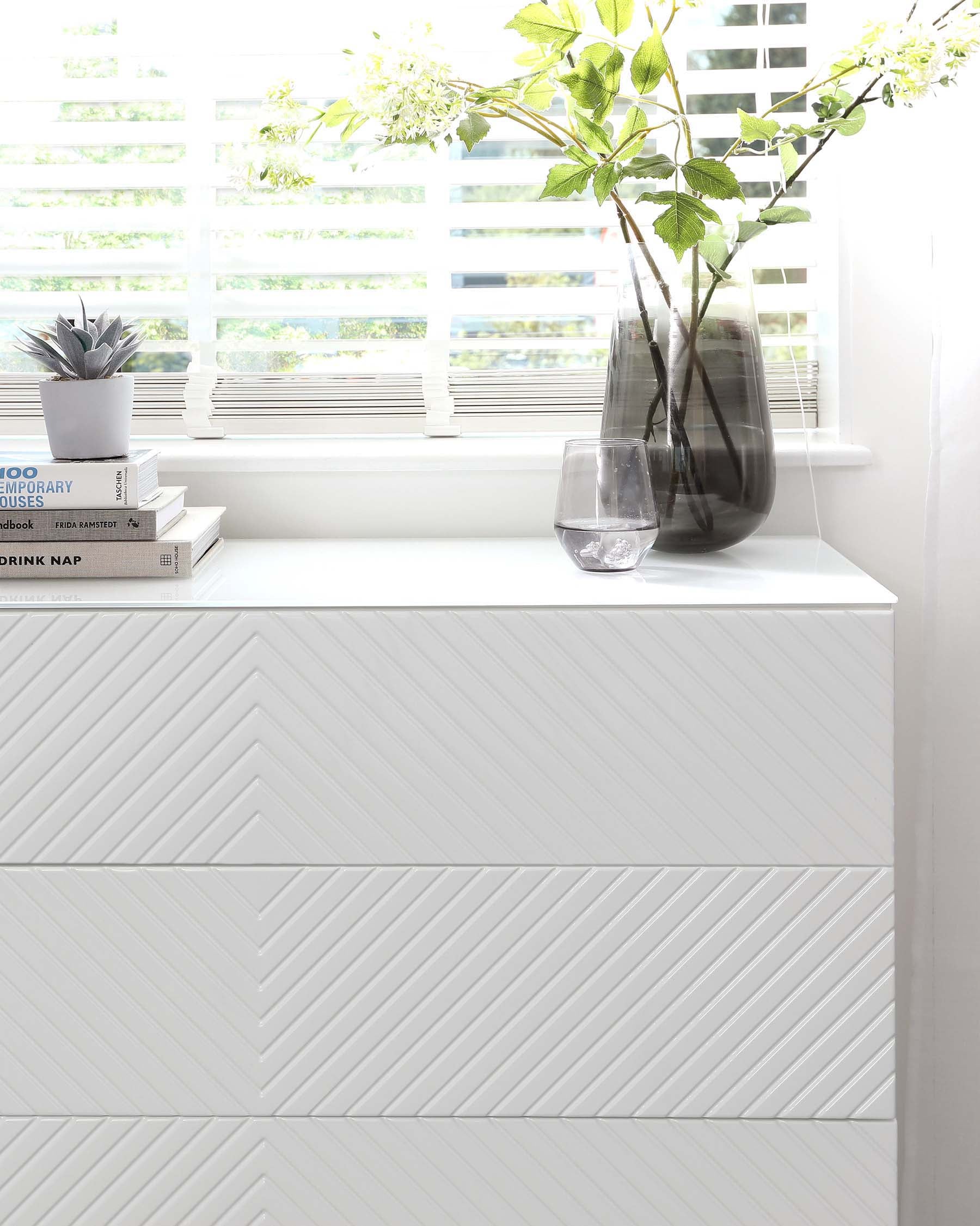 Modern white dresser with a unique chevron pattern on the drawers, placed against a wall near a window with white blinds.