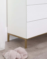 Modern white chest of drawers with a textured front design and brass-coloured metal legs, partially obscured by a soft pink fluffy pair of slippers in the foreground.