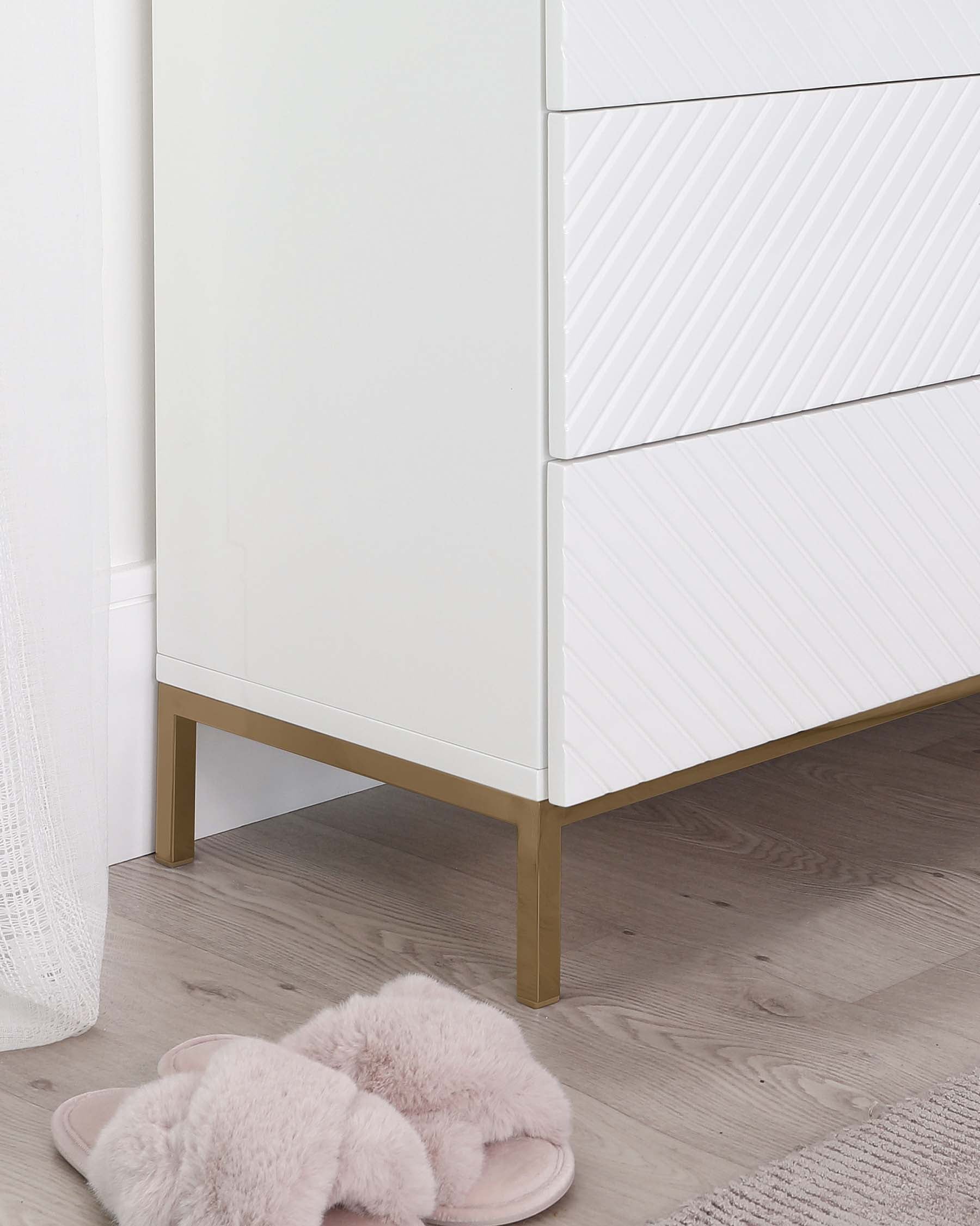 Modern white chest of drawers with a textured front design and brass-coloured metal legs, partially obscured by a soft pink fluffy pair of slippers in the foreground.