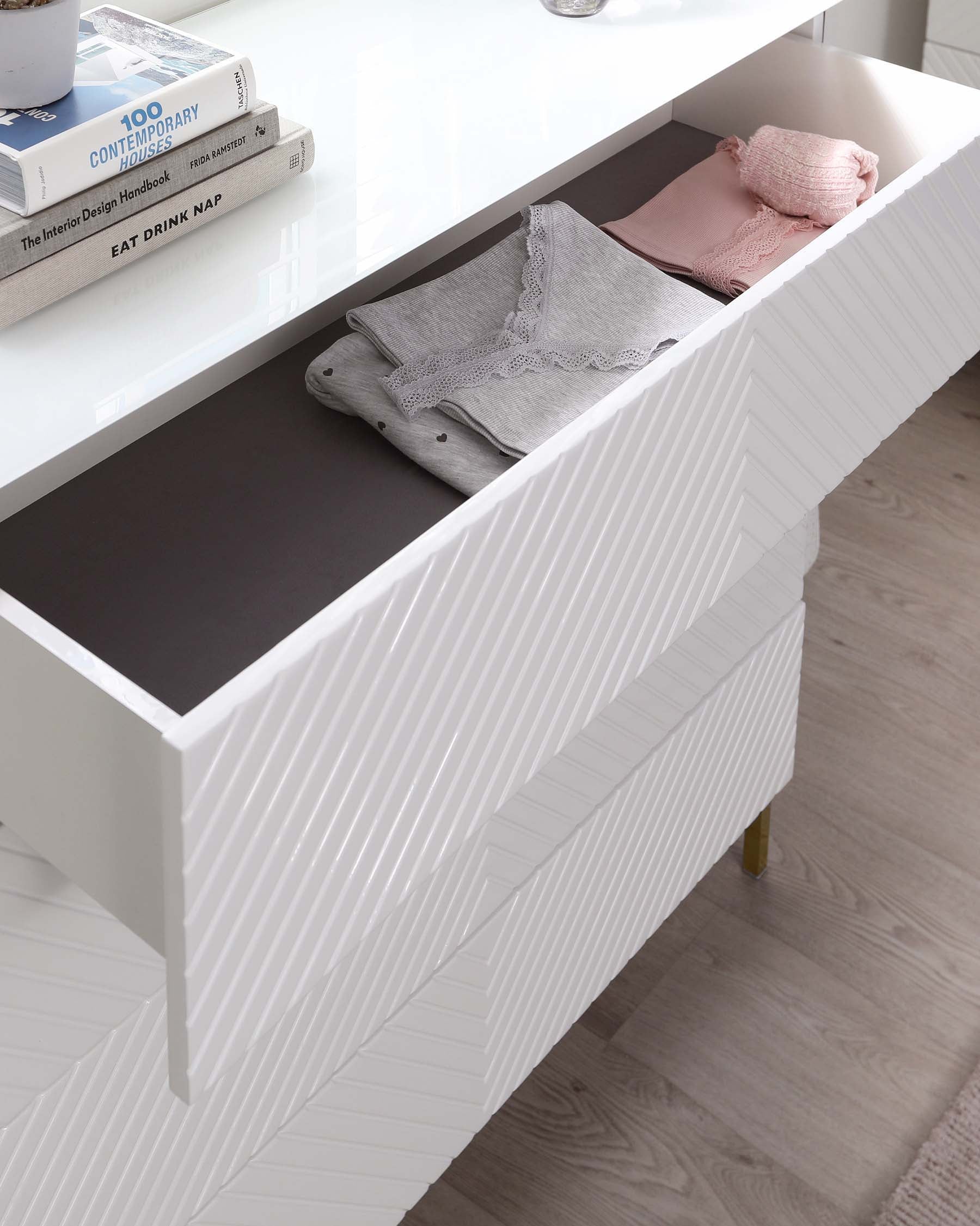 Modern white textured sideboard with an open drawer revealing neatly folded grey and pink textiles, complemented by brass capped legs and set against a light hardwood floor. A top surface of the sideboard features decorative books and a clear glass of water.