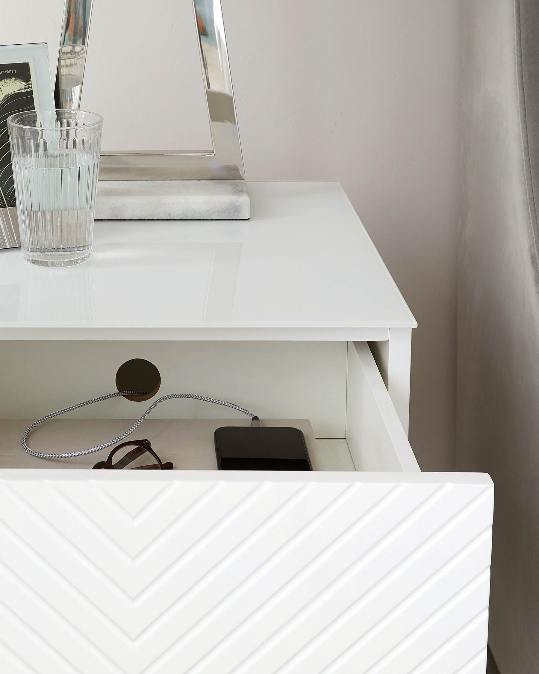 White modern minimalist vanity desk with a clean-lined silhouette and a glossy finish. Features a spacious tabletop, one open cubby shelf with charging cables, and a raised panel drawer front with a hidden drawer for additional storage.