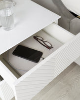 Modern white bedside table with an open drawer revealing a smartphone and eyeglasses, showcasing a ribbed design on the drawer front. A glass of water rests on the tabletop against a backdrop of a light grey upholstered bed. The scene is complemented by a light wooden floor, emphasizing a minimalist and contemporary aesthetic.