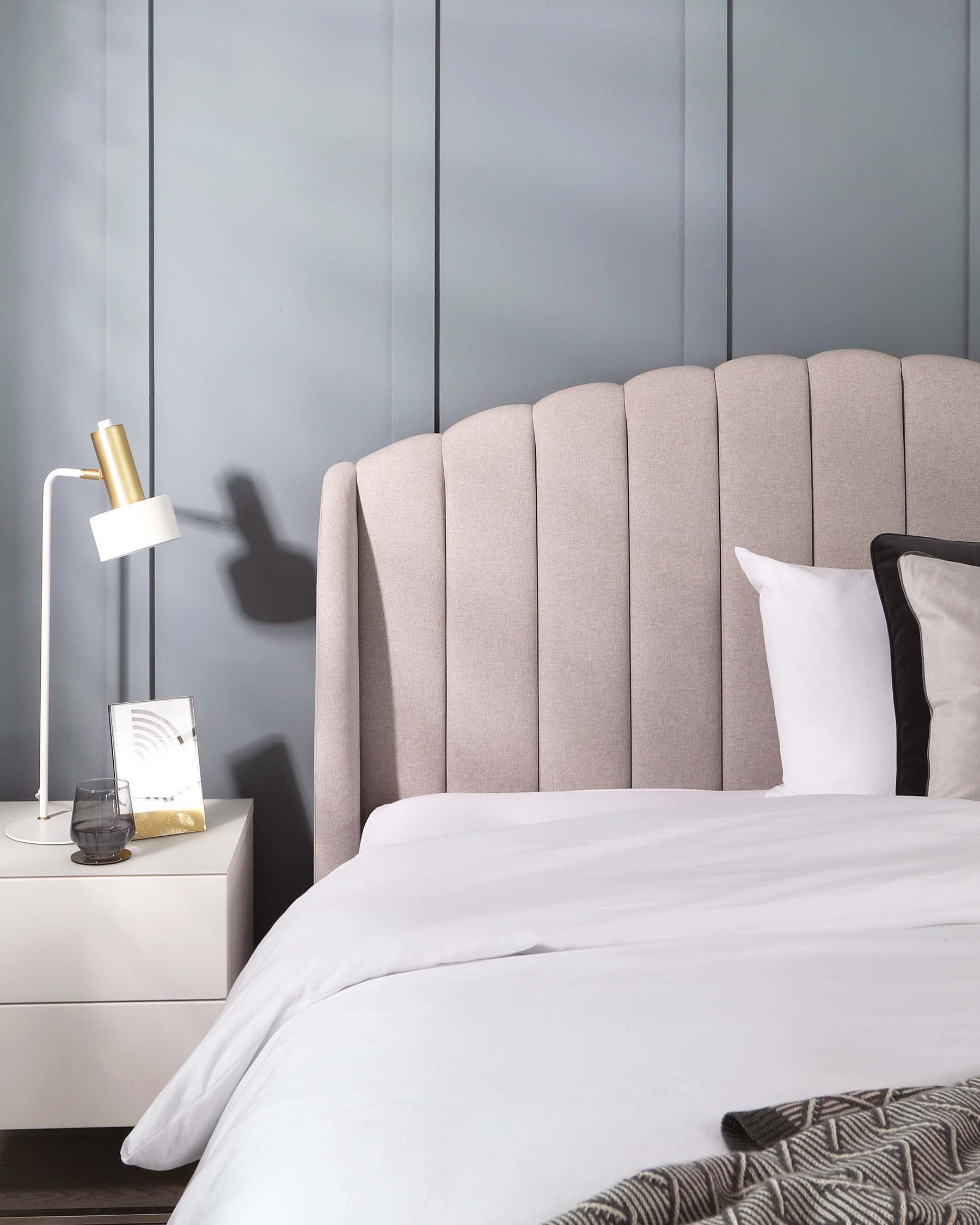 A modern bedroom showcasing a tall, beige, vertically channel-tufted headboard paired with a white bed and bedding. To the side, there's a sleek white nightstand topped with a contemporary white and gold lamp, as well as a few decorative items.