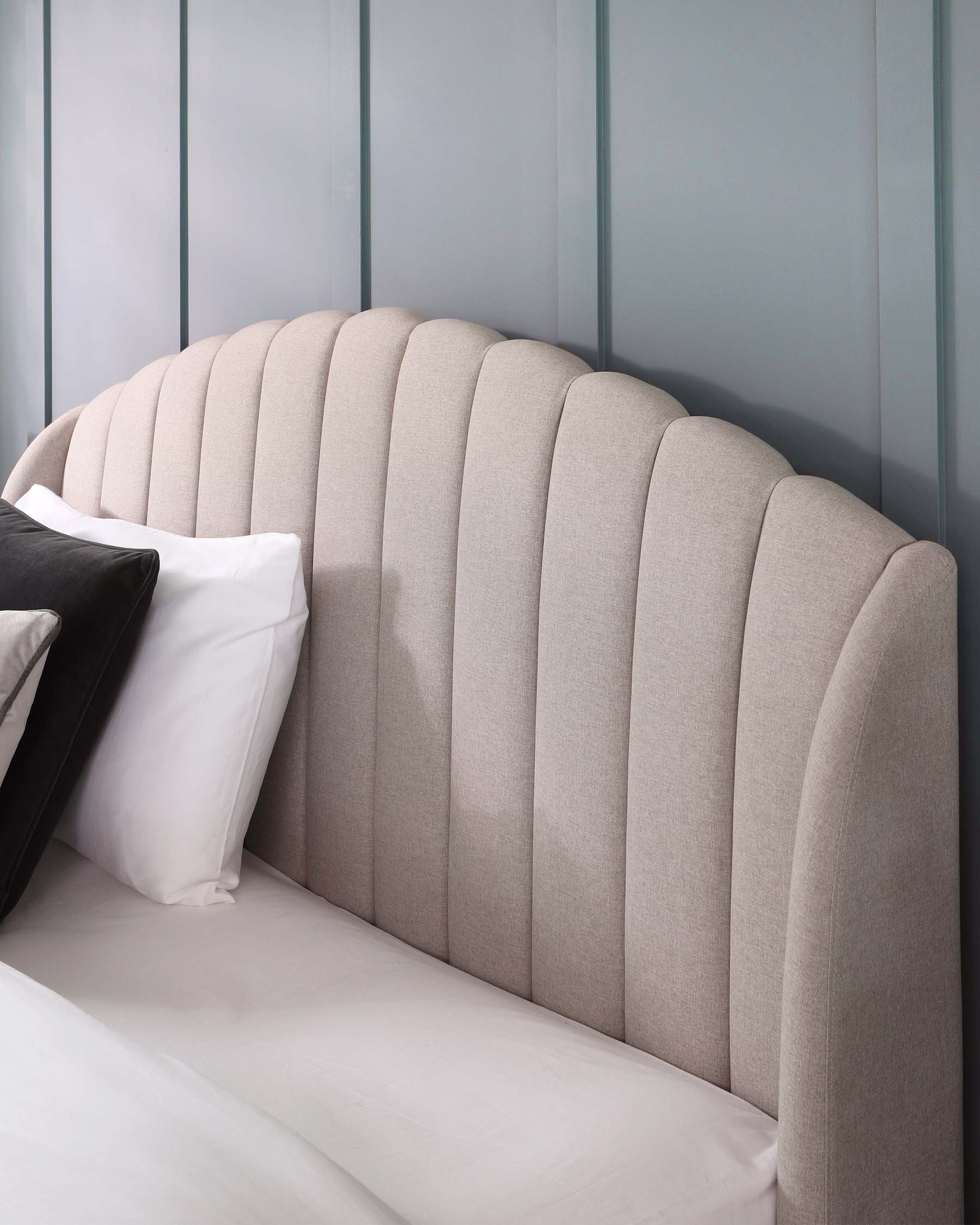 Elegant vertical channel-tufted headboard in a neutral beige fabric with a curved top design, set against a panelled grey wall, partly dressed with white and black pillows on a bed.