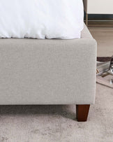 Modern upholstered bed bench in light grey fabric with four square tapered wooden legs in a dark finish, positioned at the foot of a bed with white bedding, over a textured light grey carpet.