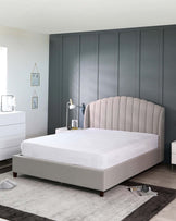 An elegant upholstered bed frame with a scalloped headboard and matching platform base in a neutral tone, accompanied by a white, minimalist nightstand featuring sleek lines and a drawer. The room also includes a grey built-in closet, with décor accents including a wall-mounted triangular shelf with framed art, a bedside lamp, and a small cluster of decorative items on the nightstand, all on a subtle grey area rug.