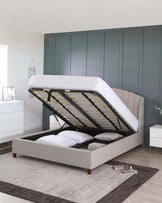 Contemporary bedroom featuring a light grey upholstered storage bed with a tufted headboard and a lifted mattress platform revealing under-bed storage. Matching white bedside tables and a dresser with clean lines and minimalist handles complement the bed. A large grey area rug anchors the space. The room's decor includes neutral wall tones and a segmented blue-grey closet system in the background, enhancing the modern aesthetic.