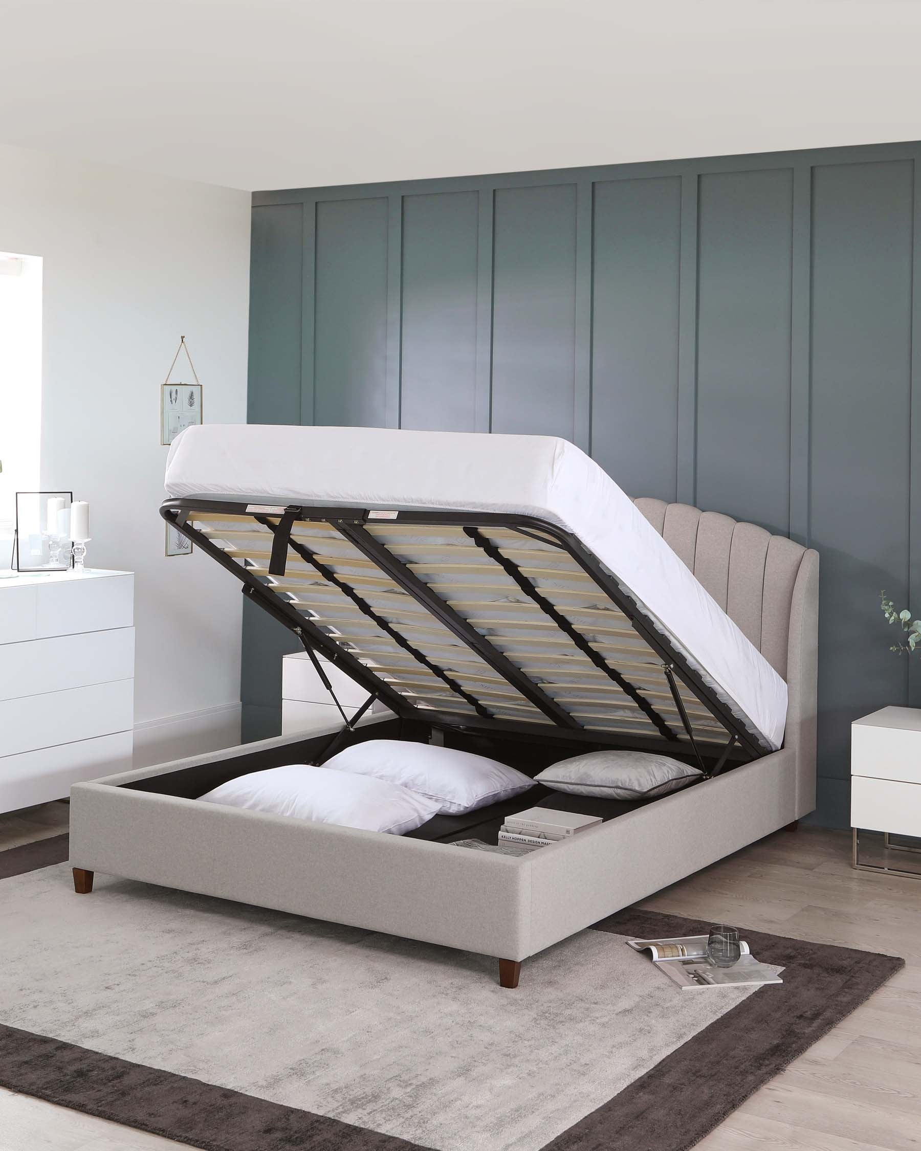 Contemporary bedroom featuring a light grey upholstered storage bed with a tufted headboard and a lifted mattress platform revealing under-bed storage. Matching white bedside tables and a dresser with clean lines and minimalist handles complement the bed. A large grey area rug anchors the space. The room's decor includes neutral wall tones and a segmented blue-grey closet system in the background, enhancing the modern aesthetic.