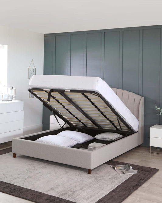 Upholstered lift-up storage bed with a tufted headboard and wood feet, featuring wooden slats partially visible under a raised mattress platform. A white nightstand with drawers and a clear glass lamp to the side; all staged on a grey area rug with a decorative clock and unlit candle as accents.