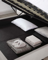 Modern storage bed with a hydraulic lift system, showcasing an upholstered frame in a dark colour with a hidden compartment beneath the lifted mattress support slats.