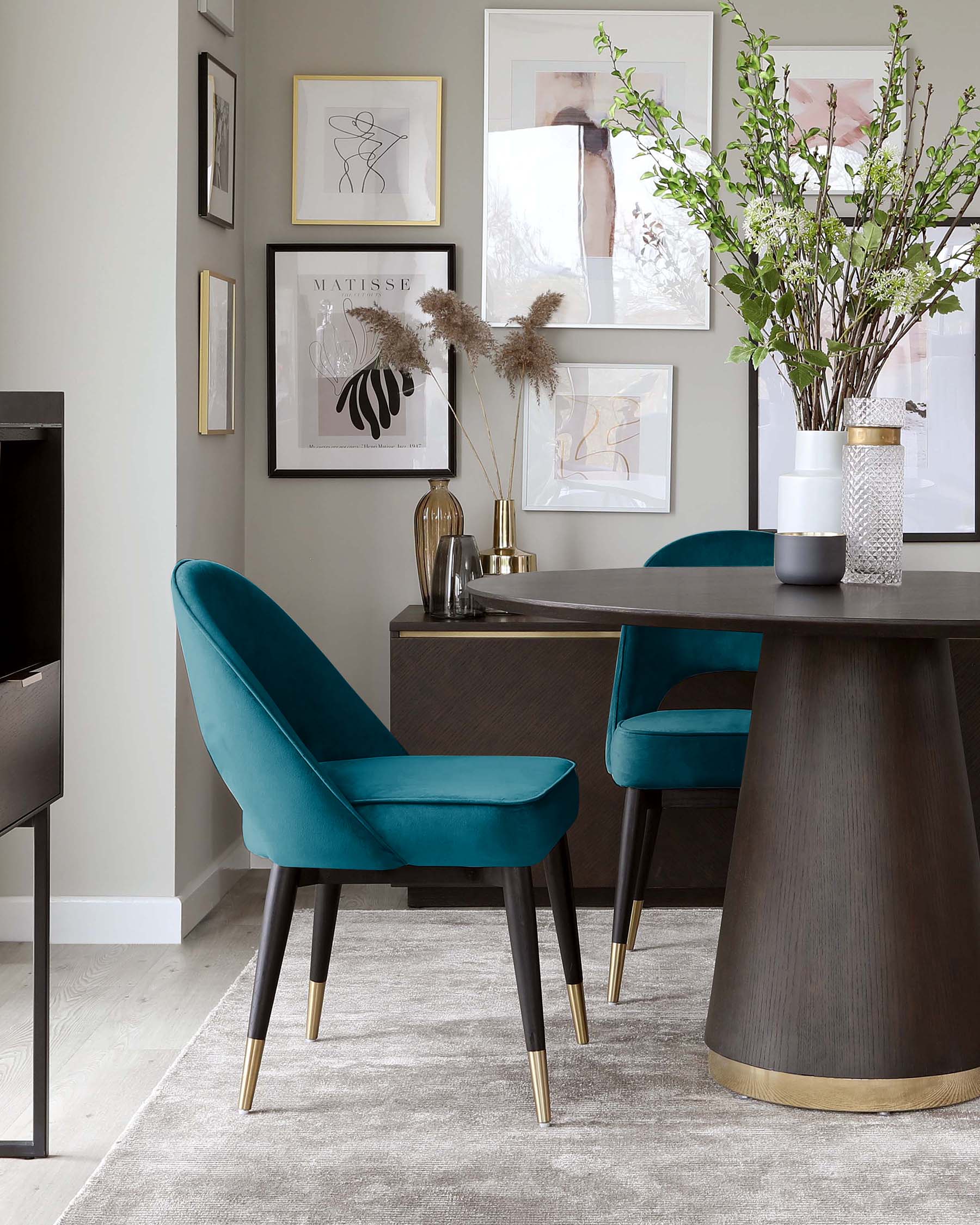 Elegant dining area featuring two plush teal blue velvet chairs with tapered black legs and gold feet caps, and a contemporary round dark wood dining table with a conical base trimmed with a gold metal band.