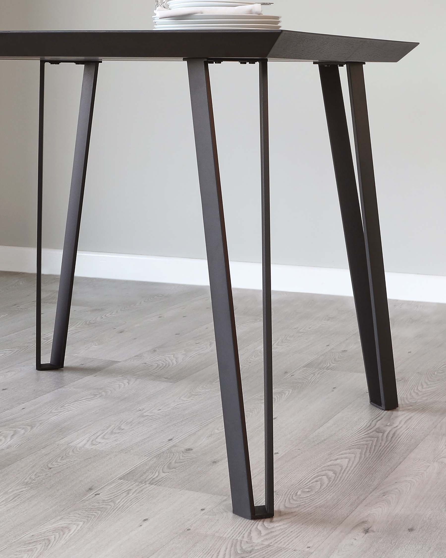 Modern minimalist black table with angled metal legs and a stack of white dishes on top, set against a light wooden floor and a white wall background.