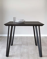 Modern minimalist dark wood dining table with sleek black angled legs, set against a neutral wall and light wooden floor. The table is adorned with a stack of white plates and a patterned glass.