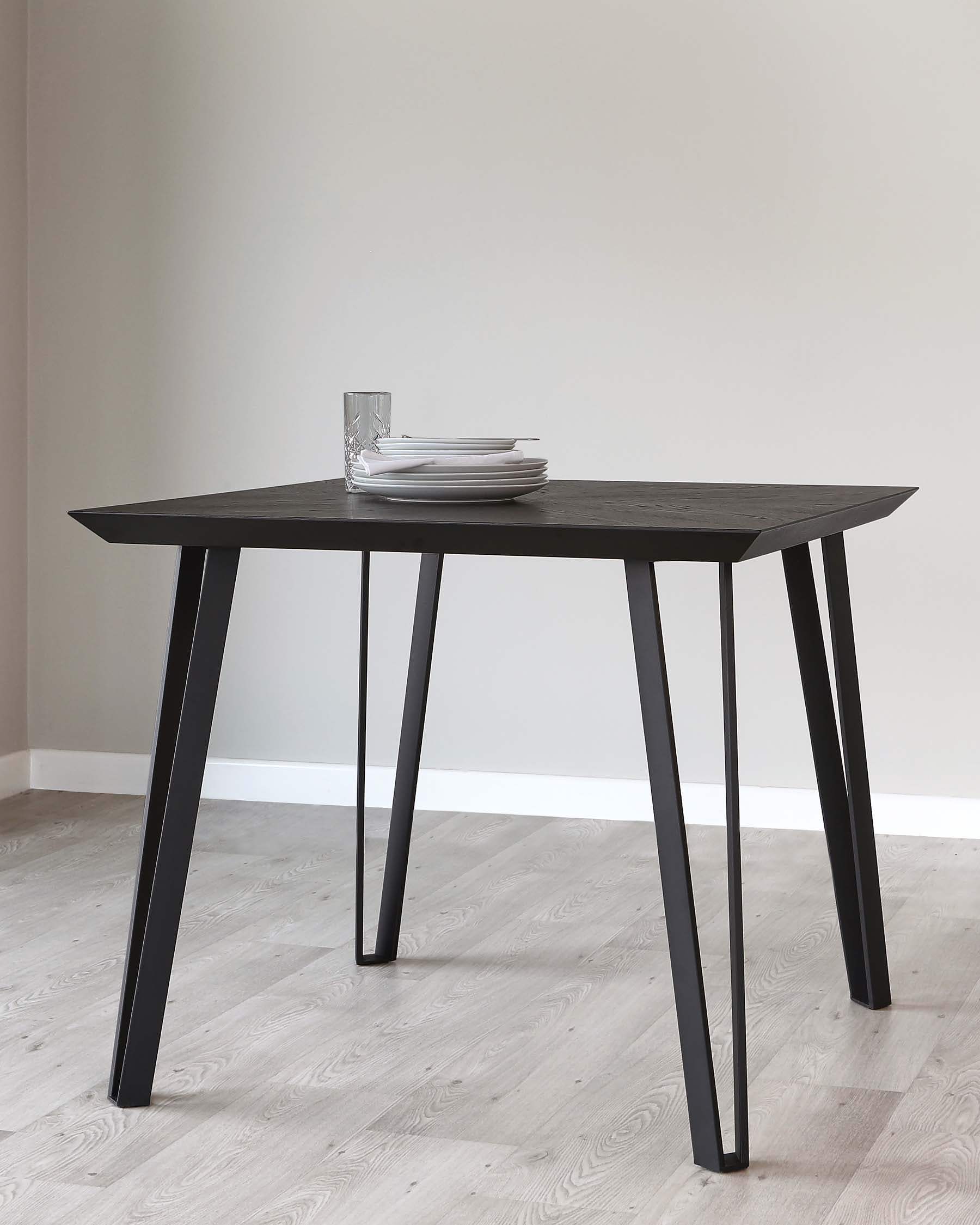 A modern, minimalist black dining table with a rectangular top and four slanted legs. On top of the table, there is a stack of white dinner plates and a clear glass. The room has light wooden flooring and soft white walls.