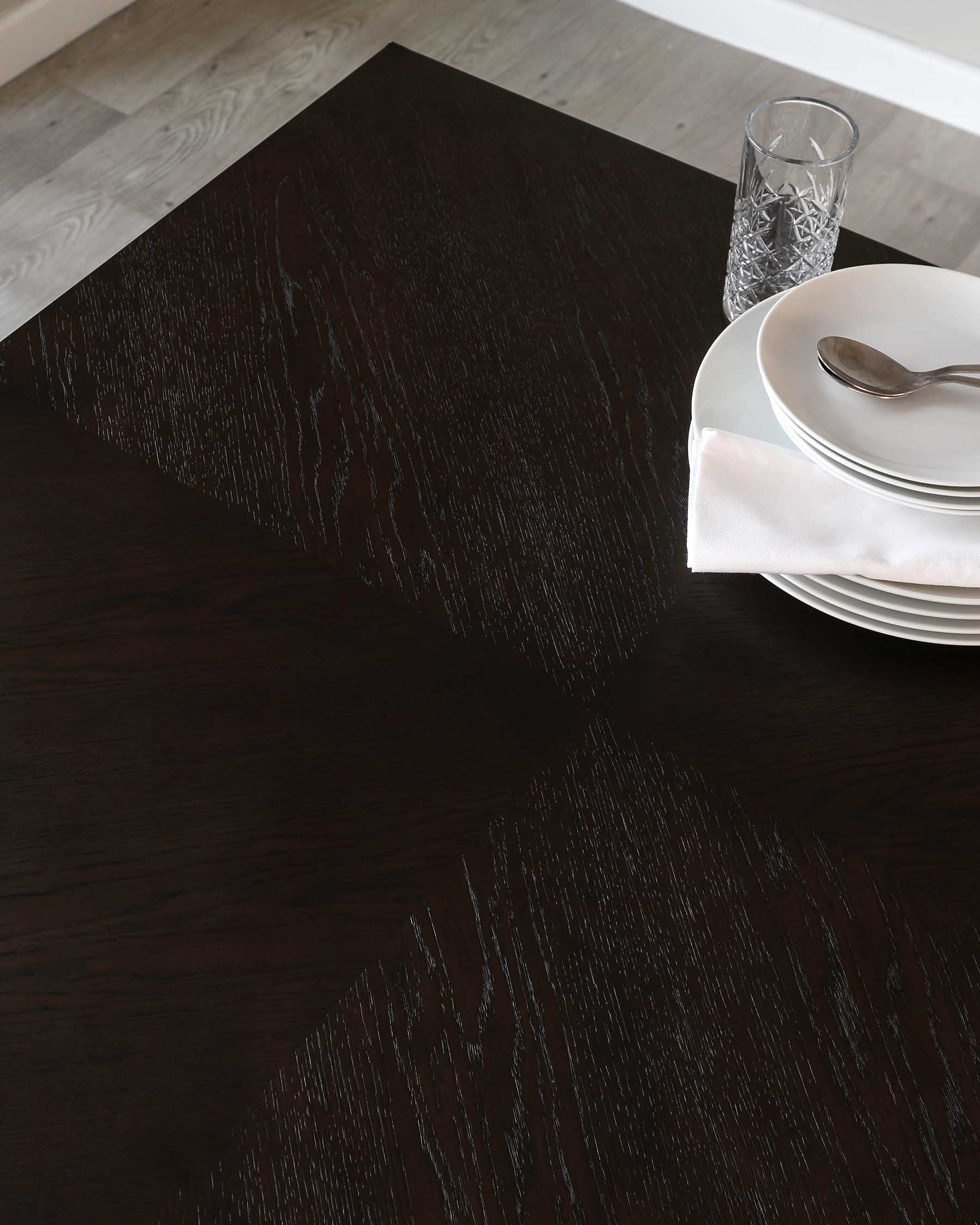 Elegant dark wood dining table with a smooth finish and visible wood grain detail, partially shown with a set of white plates, a folded napkin, and a cutlery set on top, next to a patterned glass tumbler, all suggesting a refined dining experience.