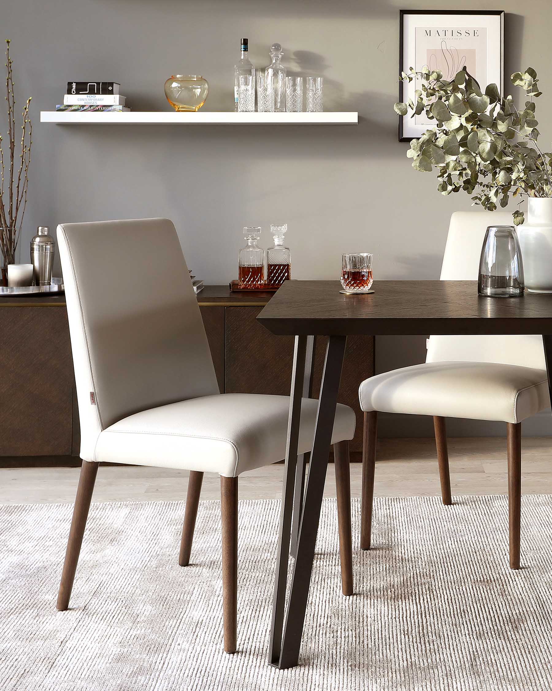 Contemporary dining set featuring a rectangular table with a rich, dark wooden top and angular black metal legs. Accompanied by three elegant, upholstered dining chairs in a light cream colour, each with slender wooden legs that complement the table's warm tones. A simple, white floating shelf is mounted on the wall above, displaying decorative items.