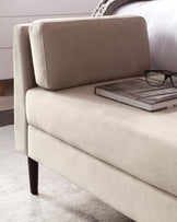 Elegant modern beige upholstered bed with a high headboard and a soft plush finish. Features straight lines and dark wood tapered legs with a book and eyeglasses resting on the edge. Part of a knitted throw blanket is visible, suggesting a cosy bedroom setting.