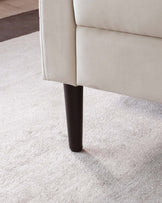 A section of a contemporary beige sofa with a close-up on a dark tapered wooden leg, set against a light beige carpet.