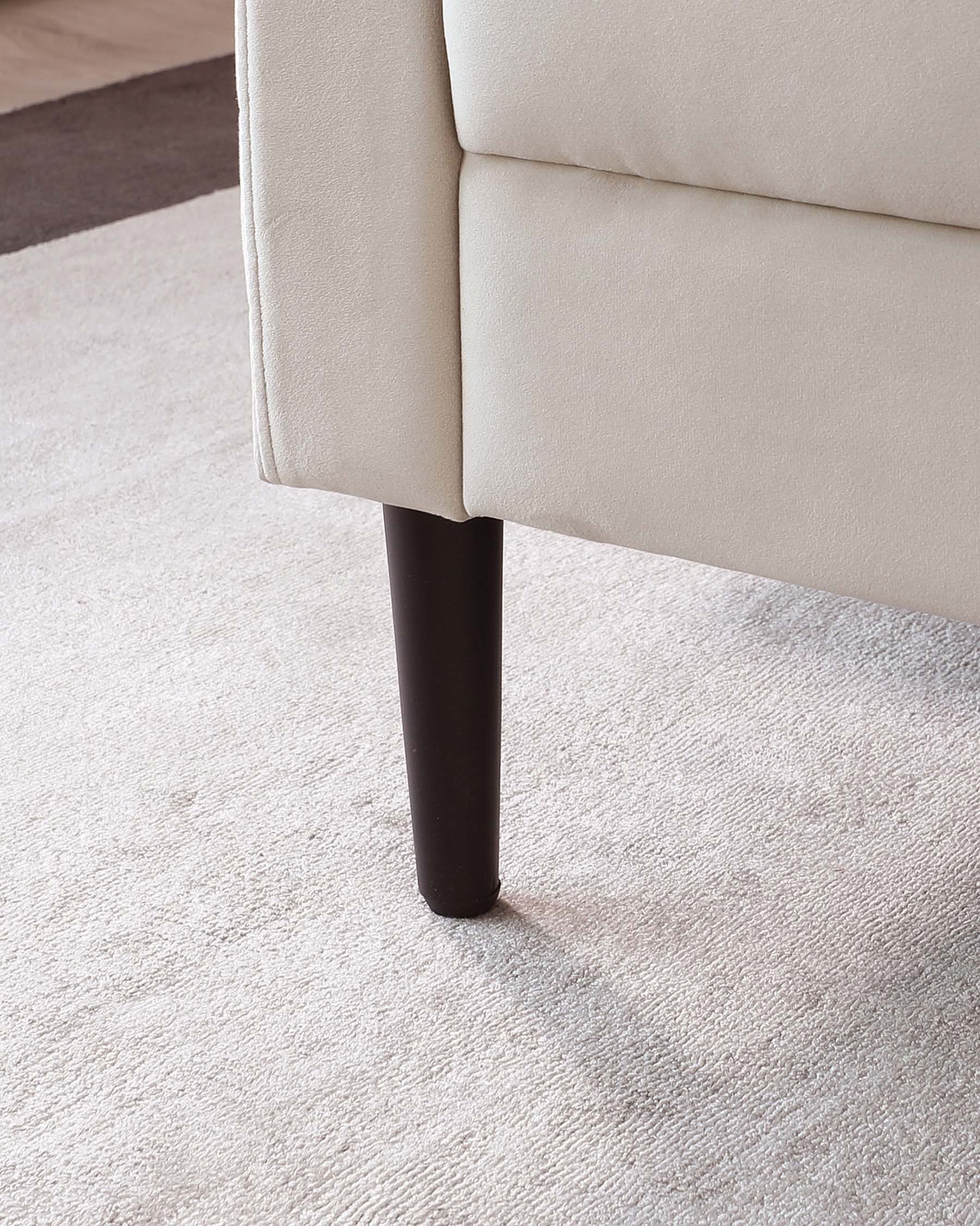 A section of a contemporary beige sofa with a close-up on a dark tapered wooden leg, set against a light beige carpet.