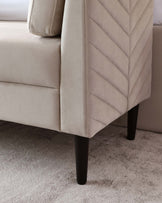 Close-up of a beige upholstered sofa with chevron patterned stitching on the sides and dark wooden tapered legs, set on a textured light grey carpet.