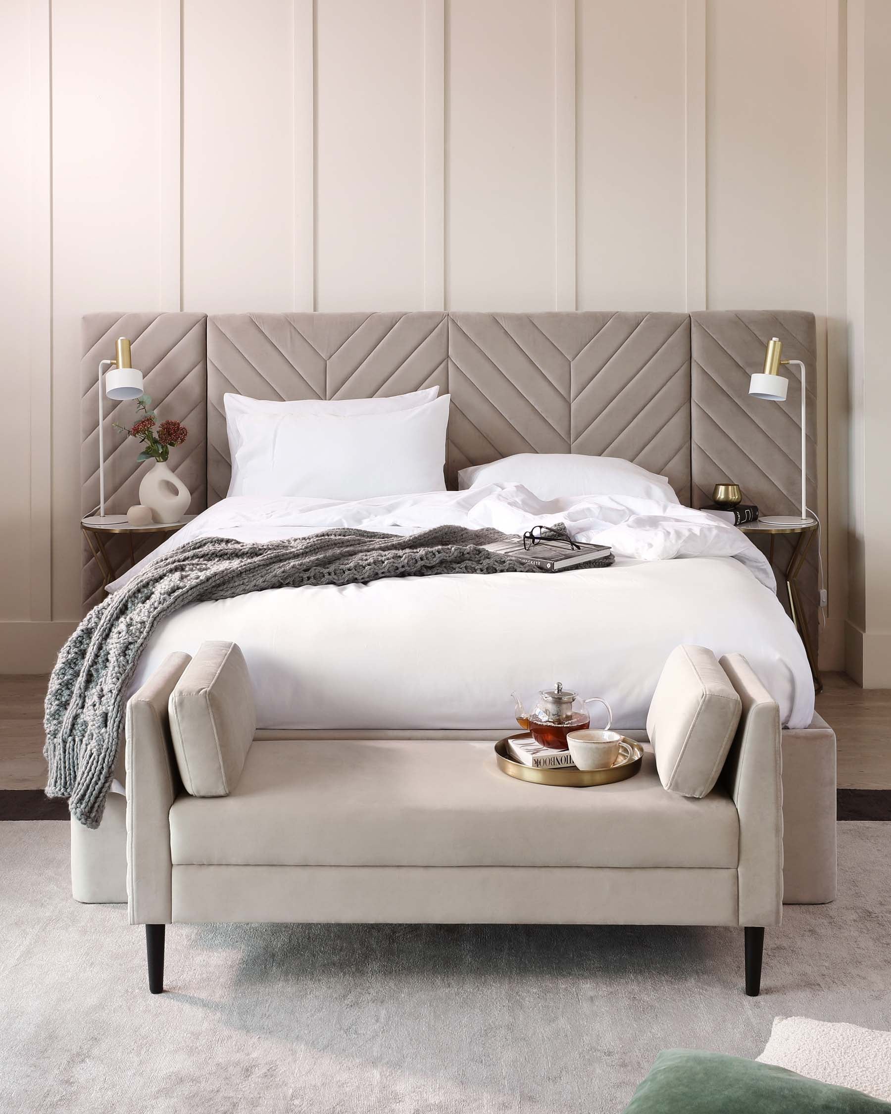 Elegant bedroom setting featuring a plush, king-size bed with a high, tufted headboard in a chevron pattern. Accompanying the bed is a soft, upholstered bench at the foot, all resting on a textured grey area rug. The scene is accented with minimalist bedside shelves and chic, adjustable reading lamps.