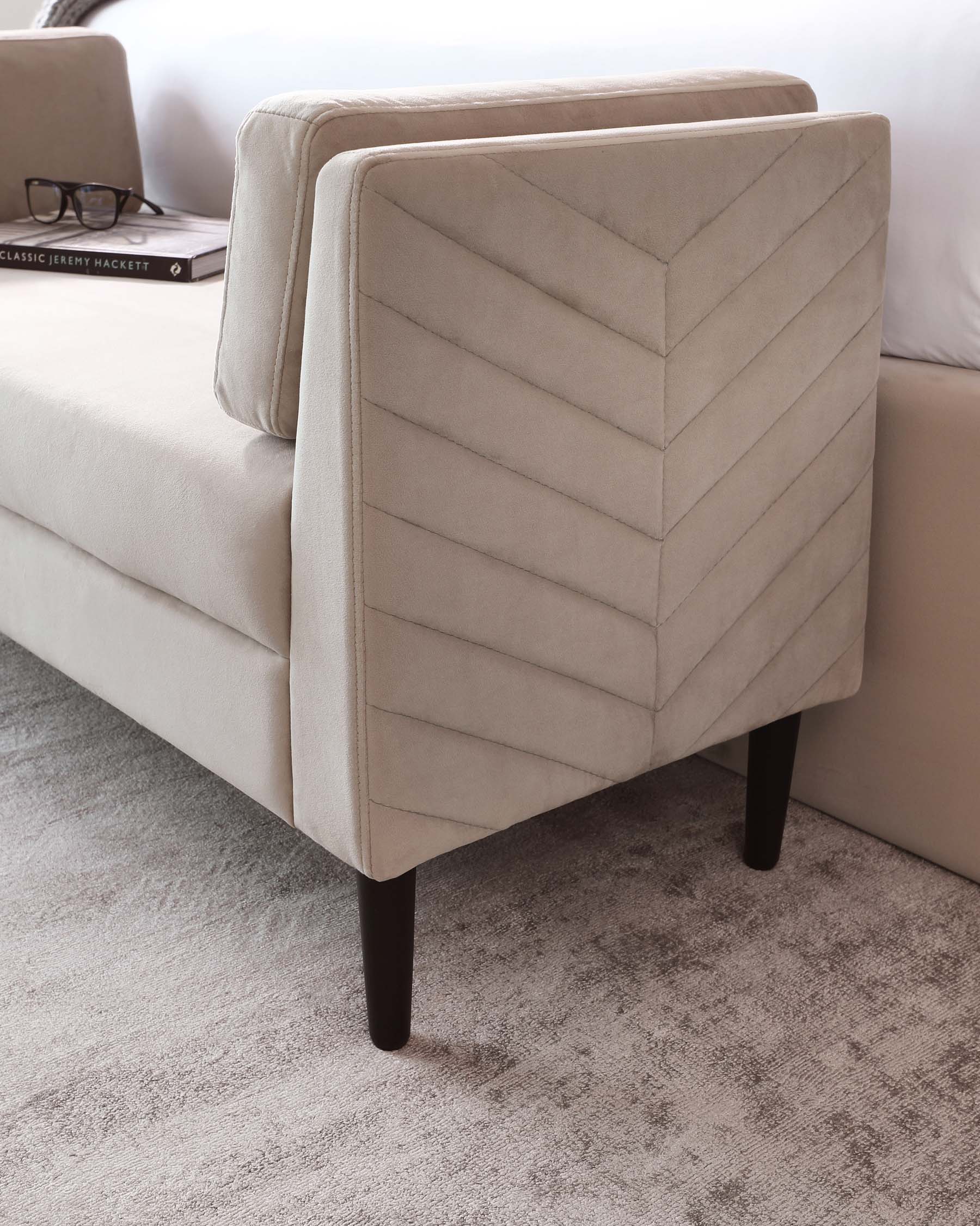 Modern beige sofa with elegant chevron upholstery pattern and dark wooden legs, partially draped by a grey knitted throw blanket, positioned on a textured grey area rug. A pair of eyeglasses and a “CLASSIC JEREMY HACKETT” book rest on the sofa, suggesting a cosy reading nook.