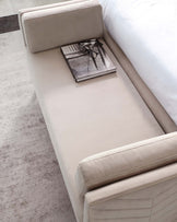 Modern beige upholstered bed bench with a minimalist design, featuring sleek lines and a smooth, cushioned seating surface, complemented by a single magazine and a pair of reading glasses resting on it.