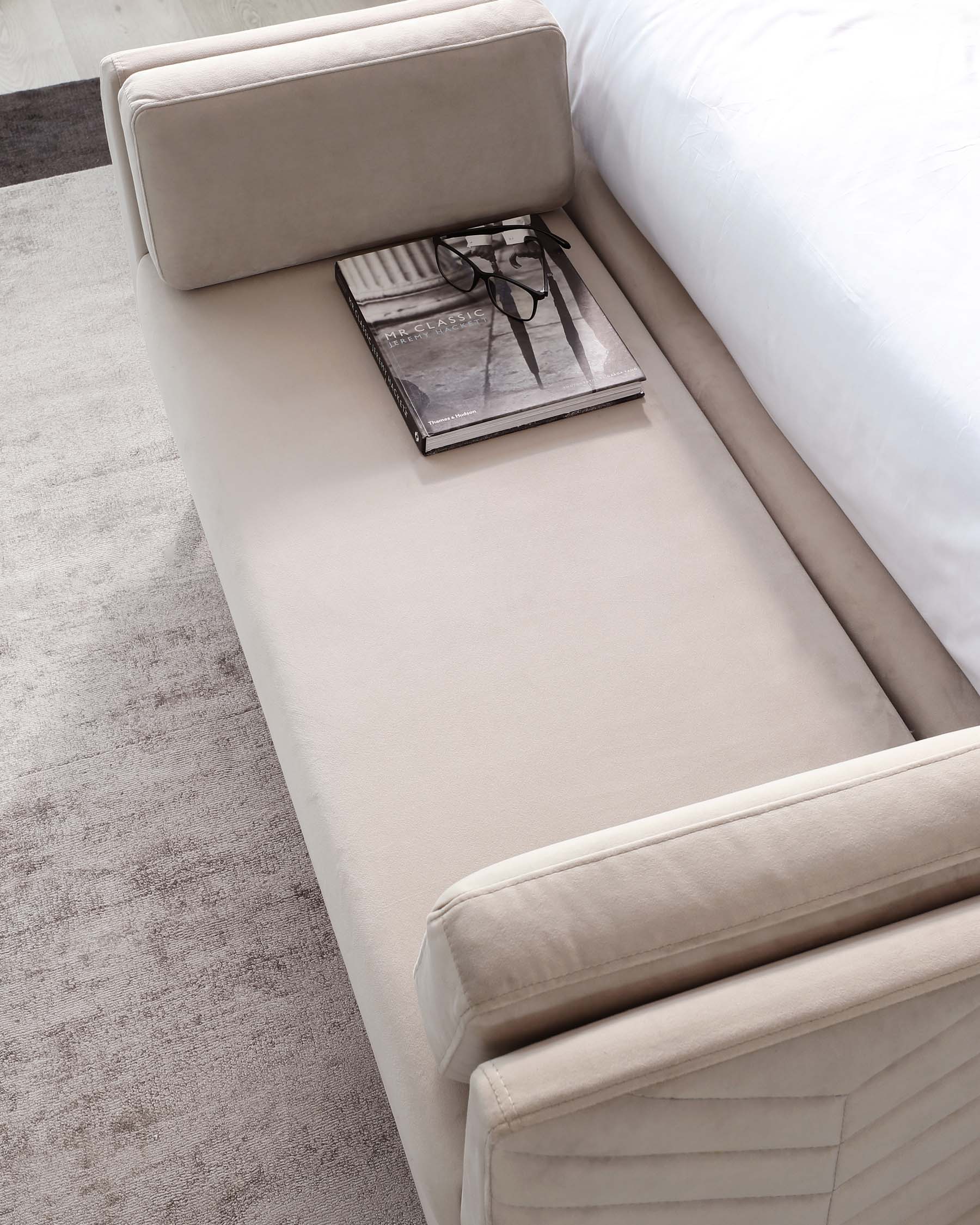 Modern beige upholstered bed bench with a minimalist design, featuring sleek lines and a smooth, cushioned seating surface, complemented by a single magazine and a pair of reading glasses resting on it.