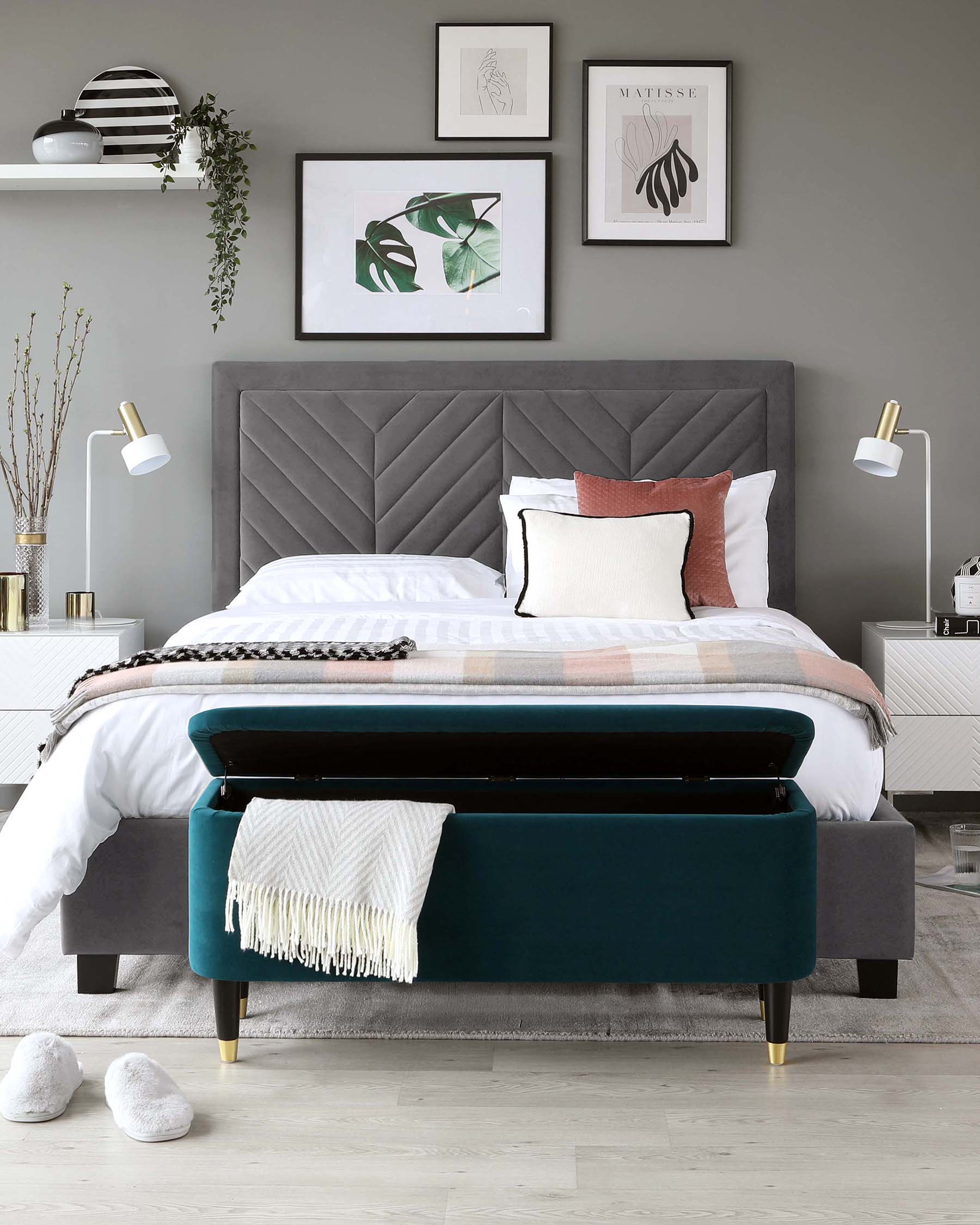 Elegant bedroom setting featuring a king-sized bed with a plush, grey, fabric-upholstered headboard featuring a diamond tufted design. In front of the bed is a luxurious, velvet upholstered bench in teal with decorative gold-tipped legs, adorned with a white fringed throw blanket resting casually on its surface. The room is styled with soft bedding, accent pillows, and ambient wall-mounted light fixtures on either side of the bed, creating a sophisticated and contemporary sleeping area.