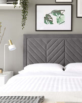 Contemporary bedroom featuring a large upholstered headboard with a geometric tufted design in a plush grey fabric. The bed is dressed in crisp white linens with subtle stripe detailing, complemented by a herringbone throw at the foot of the bed. To the side, there's a minimalist white nightstand with a sleek gold accent on the drawer handle, paired with a modern white and gold table lamp.